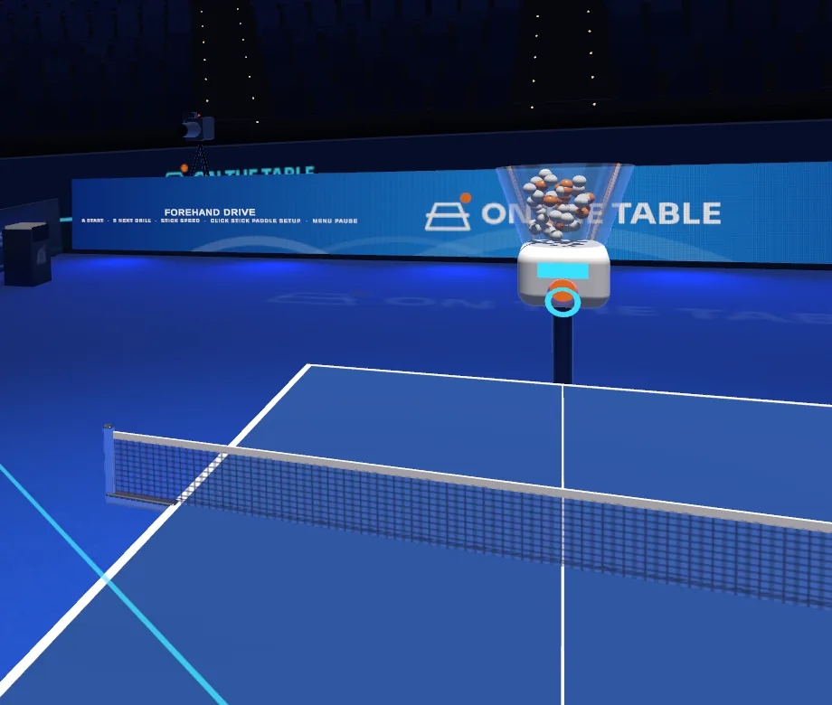 The ball machine at the far end of the table, its clear hopper full of orange and white balls, with the drill name on the LED board behind it.