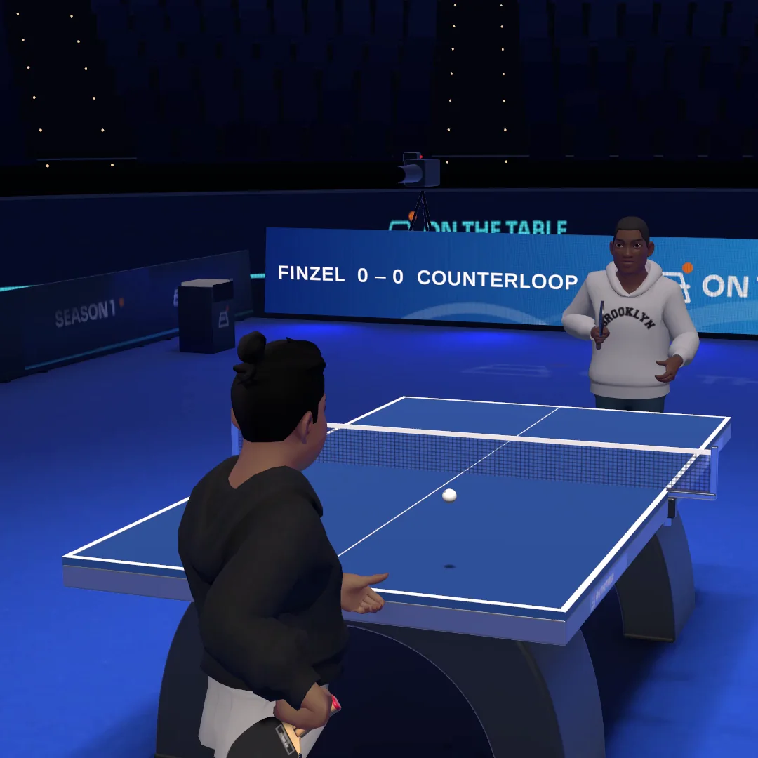 A player in a black hoodie about to serve, facing an opponent across the table in the On The Table arena, under an LED board showing 0–0.